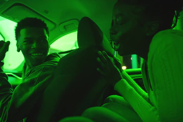 A young Black couple talking and smiling in a car at night, illuminated by colorful, changing neon lights