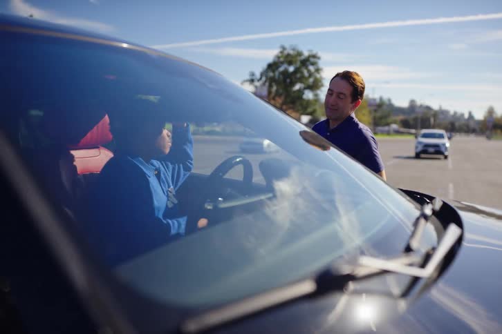 A patient father teaches his teenage son how to drive during a lesson in an empty parking lot on a sunny day