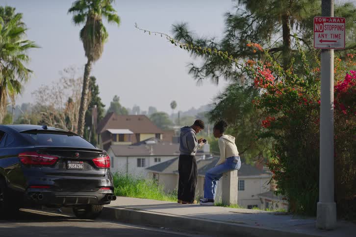 A young man takes a photo of his female friend with his smartphone as they hang out on a sunny, residential street corner