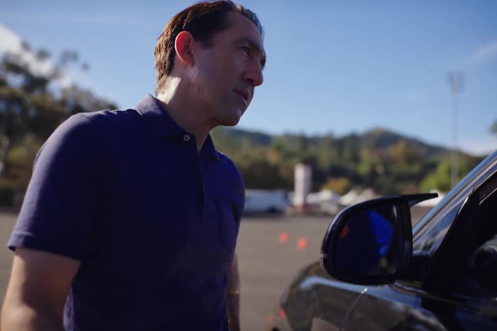 A driving instructor observes his teenage student in the driver's seat of a car in a parking lot on a sunny day