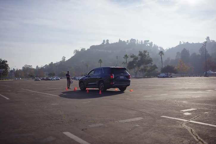 A man approaches a black SUV for a driving test in an empty parking lot with orange traffic cones