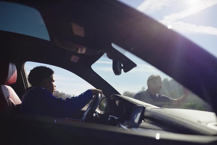 A driving instructor gives a lesson to a teenage boy in a car, viewed from inside the vehicle on a sunny day