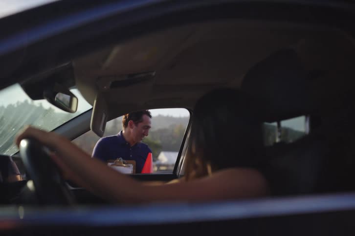 A friendly driving instructor with a clipboard and traffic cone speaks to a student driver during a lesson