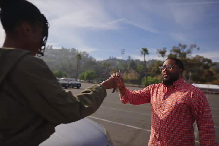 A proud father gifts his teenage daughter her first car, handing her the keys in a sunny parking lot in slow motion
