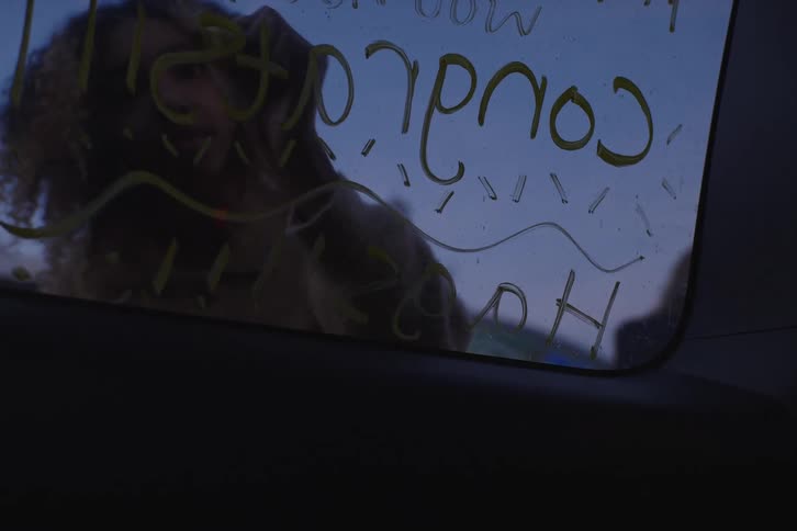View from inside a car as a young woman writes a celebratory message with a yellow marker on the window during twilight