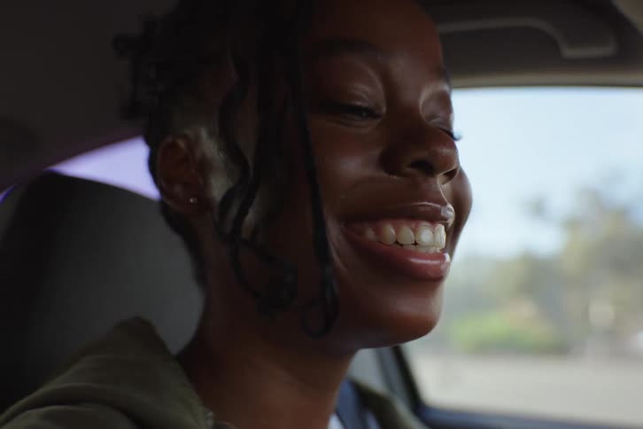 A joyful young Black woman laughs and smiles while riding in the passenger seat of a car on a sunny day