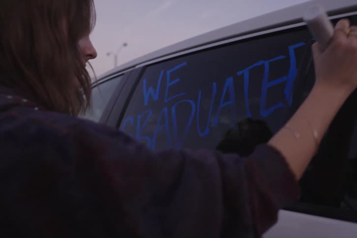 A young woman writes "We Graduated" in blue ink on a car window with a marker to celebrate