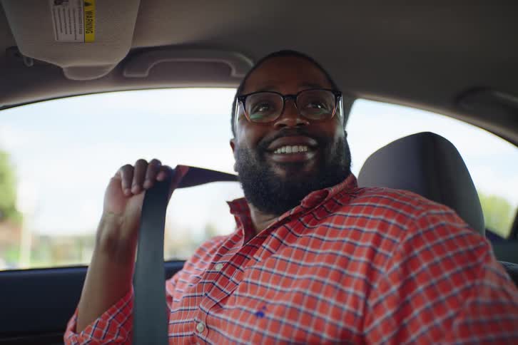 A man with a beard and glasses sits in the driver's seat, talks to someone off-camera, and then puts on his seatbelt