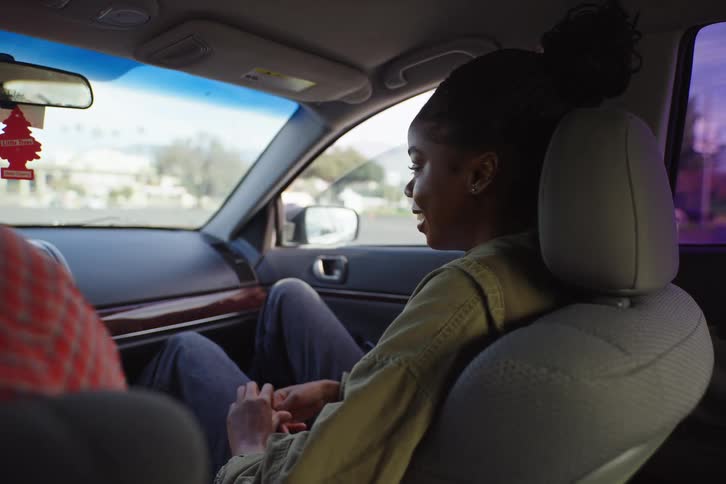 A young African American woman smiles and talks with the driver while sitting in the passenger seat of a car