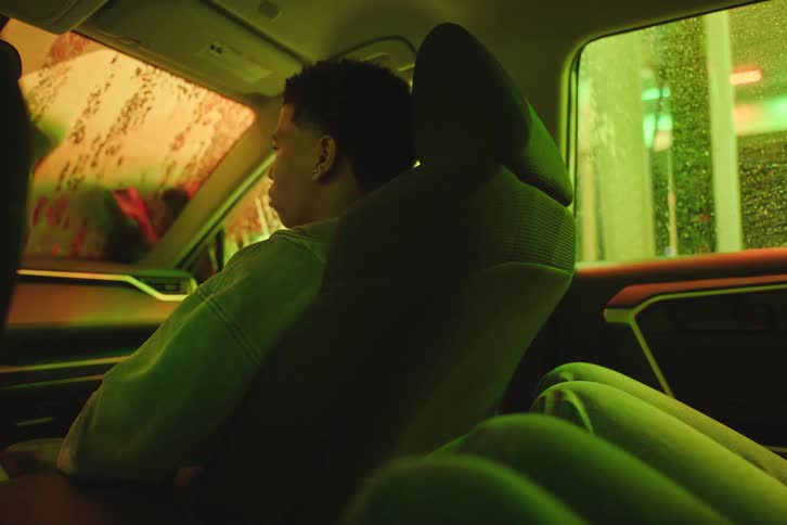 A young man sits thoughtfully in the passenger seat of a car as it moves through an automatic car wash with colorful flashing lights