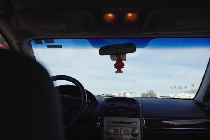 View from the backseat as a man exits a parked car and hands the keys to a smiling woman in a sunny parking lot