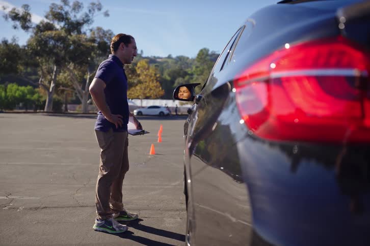 A driving instructor talks to his student, who is reflected in the side mirror, while he holds a clipboard during a driving test in a sunny parking lot