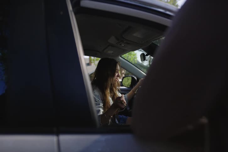 A young woman seen through a car window engages in an animated and expressive conversation while in the driver's seat