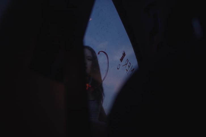 View from inside a car as a young woman draws hearts, crowns, and messages on the rear window at dusk