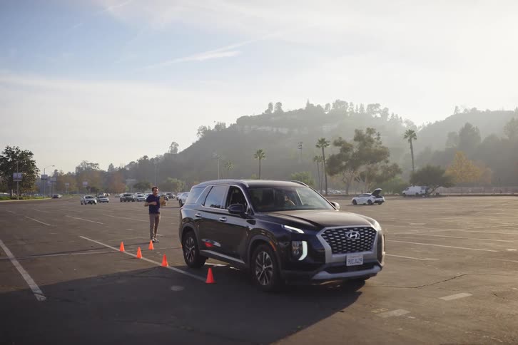 A driving instructor guides a student in a black SUV through an orange cone course in a sunny parking lot in slow motion