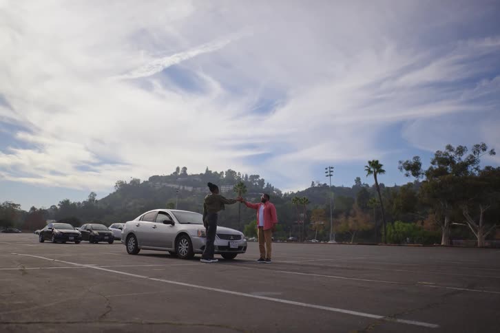 A man and woman get out of their broken-down car, fist bump, and work together to push it in a large parking lot