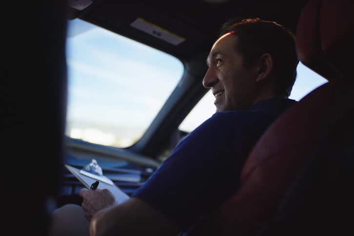 A man, possibly a coach or supervisor, smiles while observing from the passenger seat of a moving car and holding a clipboard