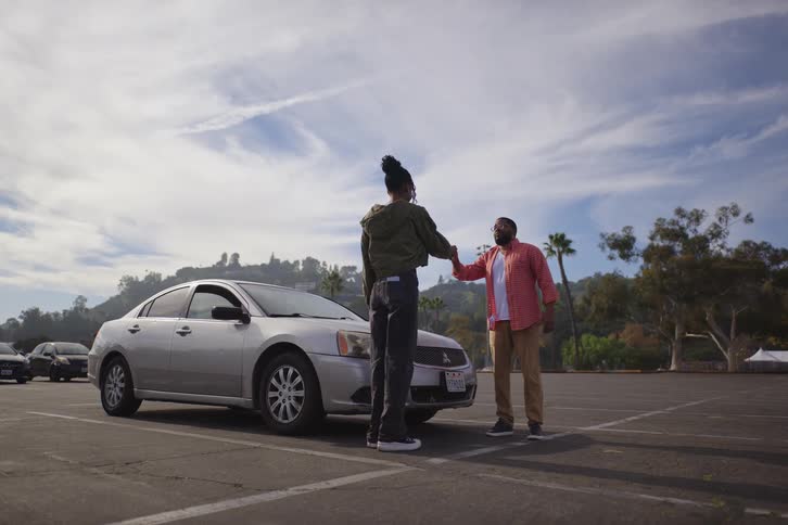 A father gets out of the driver's seat and shakes his daughter's hand before she gets in to practice driving in a large, empty parking lot