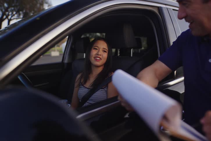 A driving instructor with a clipboard gives instructions to a young woman during her driving lesson in a parking lot