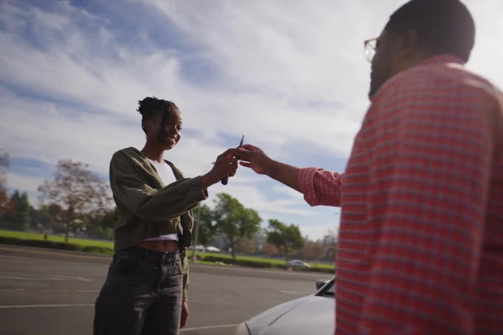 A proud father exits his car and gives the keys to his smiling daughter in a parking lot on a sunny day