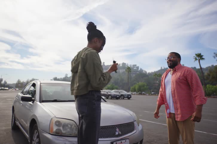 An African American father hands the car keys to his teenage daughter for a driving lesson in a sunny parking lot