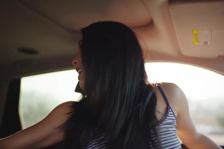 A happy young woman with long dark hair dances and sings along to music in the passenger seat of a car in slow motion