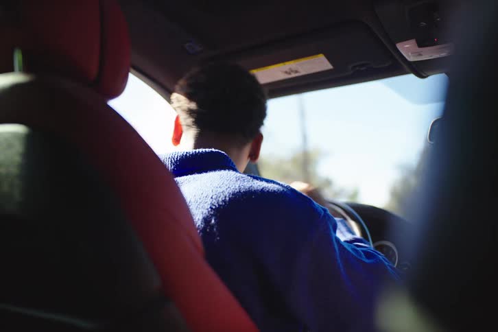 Over-the-shoulder perspective of a young man in a blue sweater driving a car on a sunny day