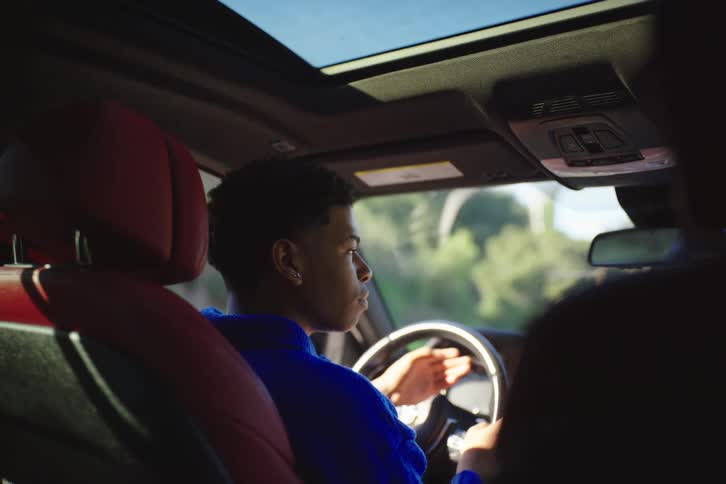 A young Black man in a blue sweater drives a luxury car, seen from the backseat on a sunny day