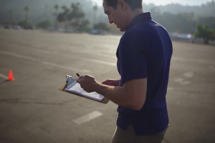 An event coordinator walks through an empty parking lot in the sun while taking notes on his clipboard