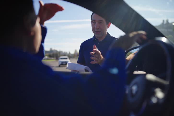 A man explains the issues with his broken-down car to a mechanic sitting inside the vehicle on a sunny day