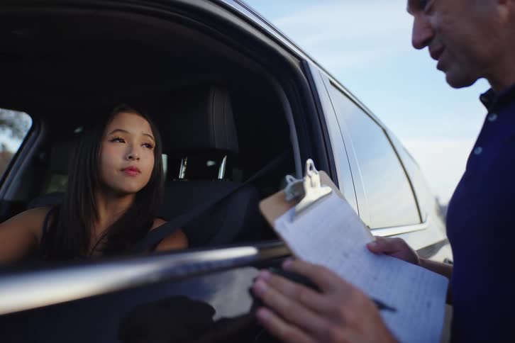 A teenage girl in the driver's seat listens intently as her driving instructor gives her a lesson from outside the car