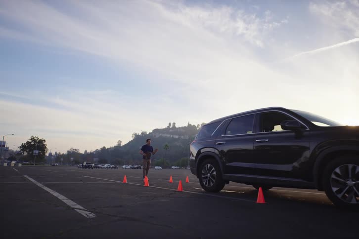 A man guides a black SUV through a driving course marked by orange cones in an empty parking lot at sunrise