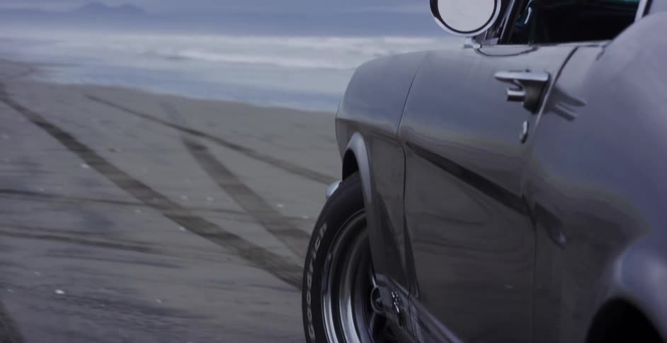A slow reveal of a classic American muscle car parked on a remote, foggy beach, starting from a close-up of its tire tracks in the sand
