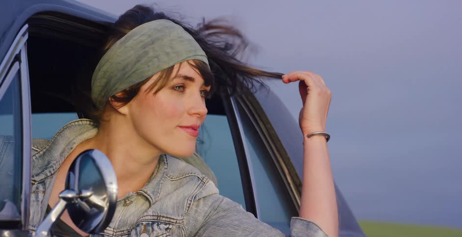 A beautiful woman in a denim jacket and headband smiles as she feels the wind in her hair from a car window