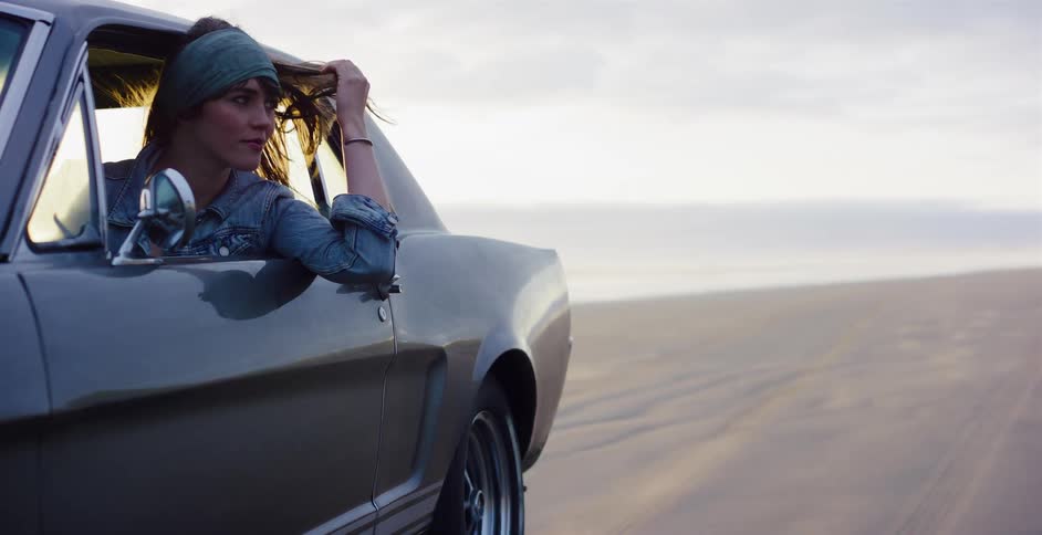 A stylish woman in a classic car enjoys the wind in her hair while parked on a sandy beach at sunset