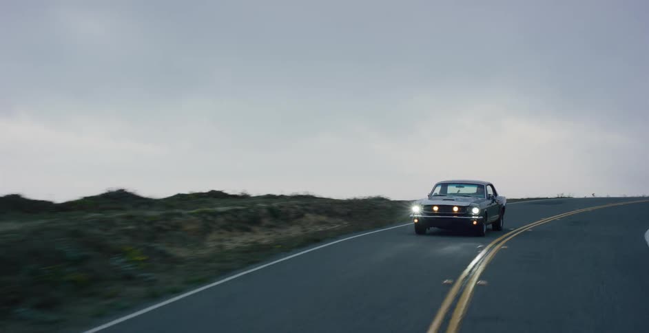 A classic muscle car drives with its headlights on down a winding coastal highway next to the ocean on a foggy, overcast day