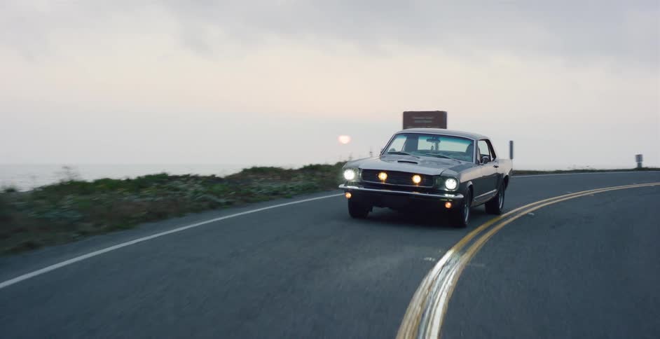 A classic grey Ford Mustang with its headlights on drives in slow motion around a bend on a scenic coastal highway at dusk