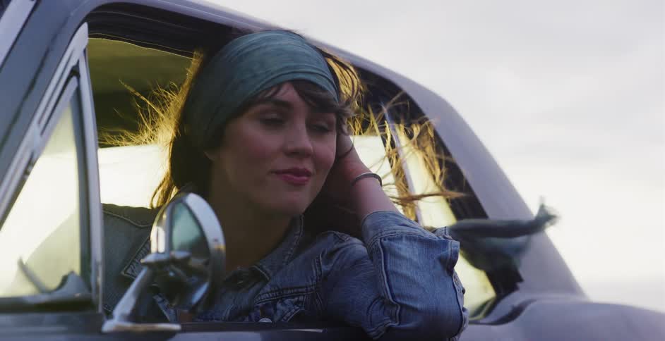 Slow motion shot of a free-spirited woman in a classic car with her hair and a scarf blowing in the breeze as she enjoys the journey at sunset