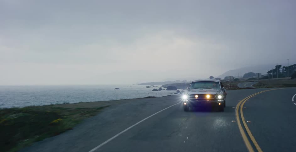 A classic muscle car drives down a winding coastal highway next to the ocean on a moody, overcast day
