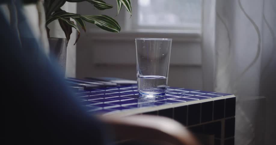 A still glass of water sits on a blue tiled table in a timelapse showing the passage of time through shifting light and reflections