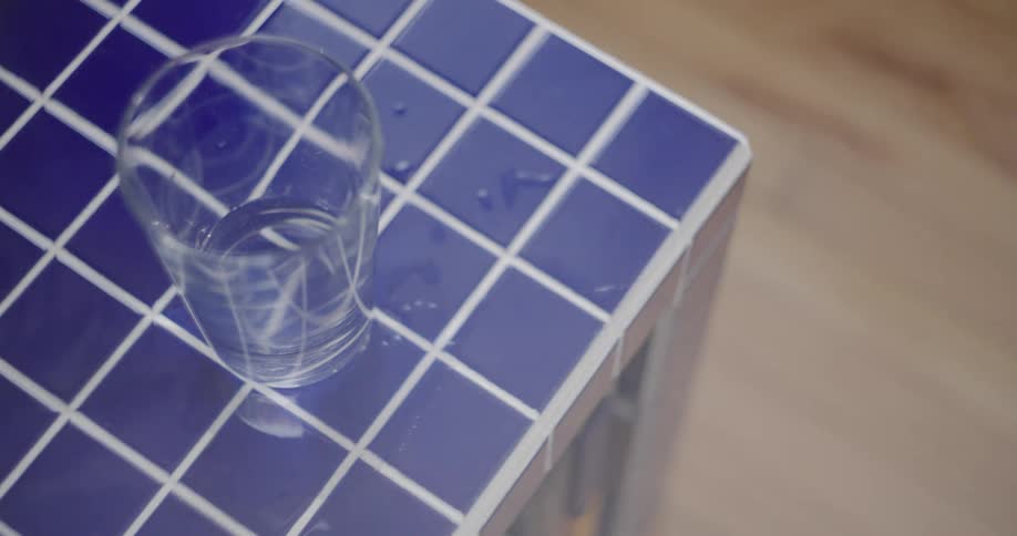 A still close-up of a glass of water resting on a blue tiled table with a passing reflection