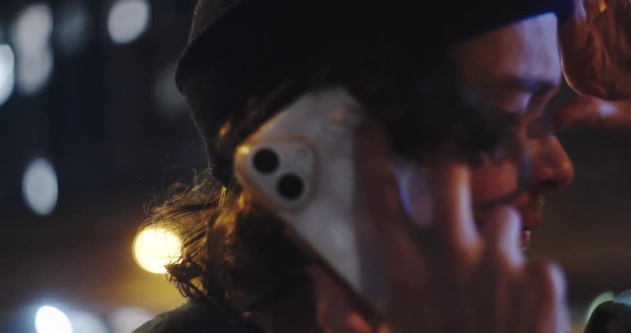 Close-up of a serious young man in a beanie talking on his smartphone against a backdrop of city lights at night