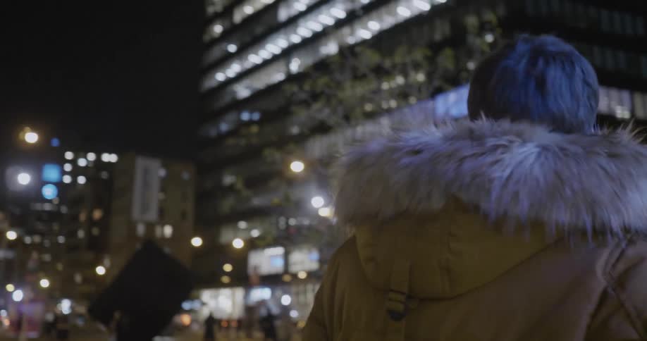 A man wearing a parka walks through a busy city square at night