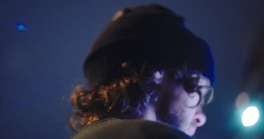 A pensive young man in a beanie looks down thoughtfully in a moody, blue-lit setting