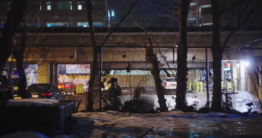 A solitary person in a beanie sits by a chain-link fence in a snowy, urban setting at night before walking away