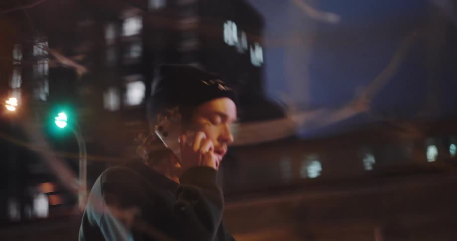 A young man with curly hair wearing a beanie thoughtfully scrolls on his smartphone before making a phone call on a city street at night