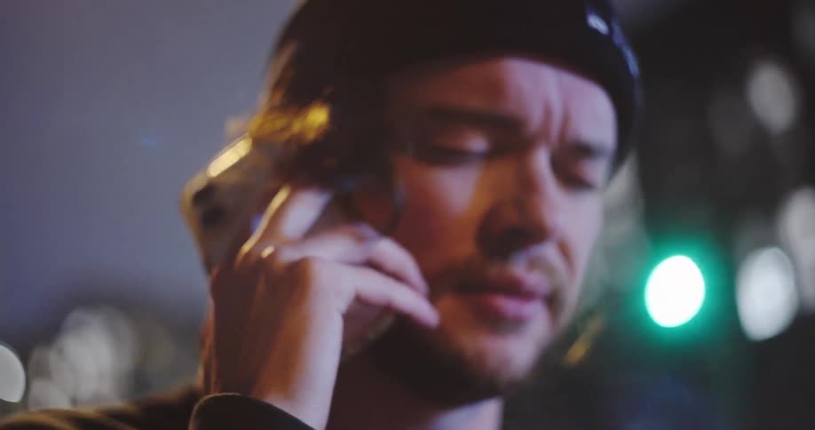 A concerned man in a beanie has an emotional conversation on his smartphone on a city street at night in slow motion