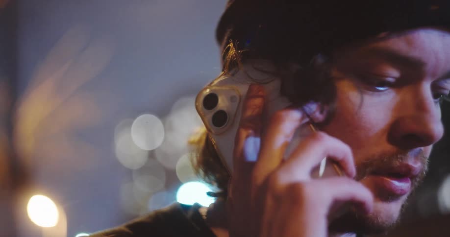 Close-up of a serious young man in a beanie talking on his smartphone on a city street at night