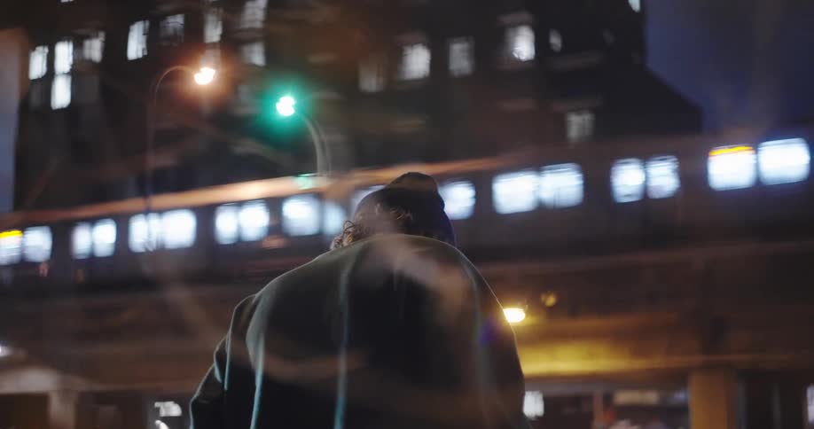 A man in a beanie walking thoughtfully as an elevated train passes through a city at night in slow motion
