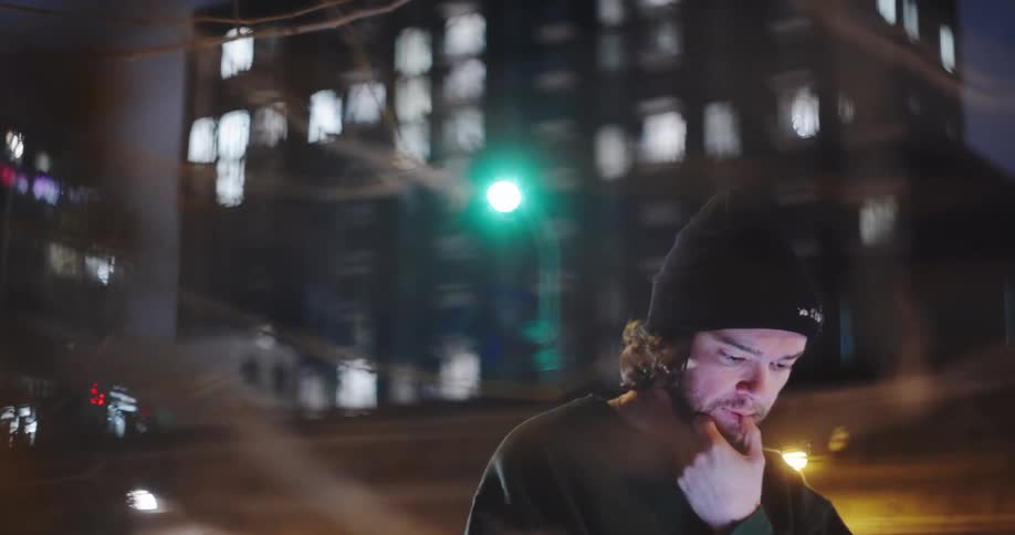 Moody cinematic shot of a thoughtful young man talking on his phone at night with an elevated train and city lights in the background